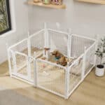 White octagonal metal dog playpen indoors with a golden retriever, pet feeder, toys, and bed in a bright room with wooden floors and a potted plant.