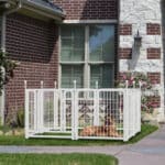 White rectangular outdoor dog playpen on grass with a German Shepherd, next to a brick-and-stone house with windows, pink flower bushes, and a paved walkway.