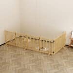 Light wood six-panel rectangular dog playpen with vertical slats, two small dogs inside, in a bright room with minimal décor.