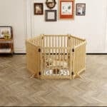 Light wood hexagonal dog playpen with vertical slats, dog resting on mat, in bright room with wall art and wooden table.