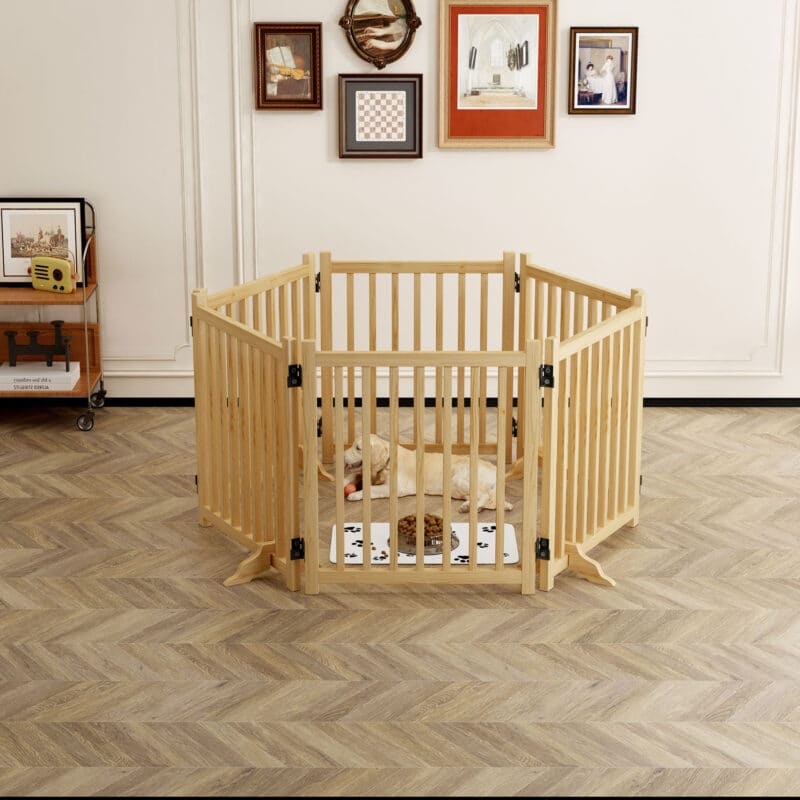 Light wood hexagonal dog playpen with vertical slats, dog resting on mat, in bright room with wall art and wooden table.