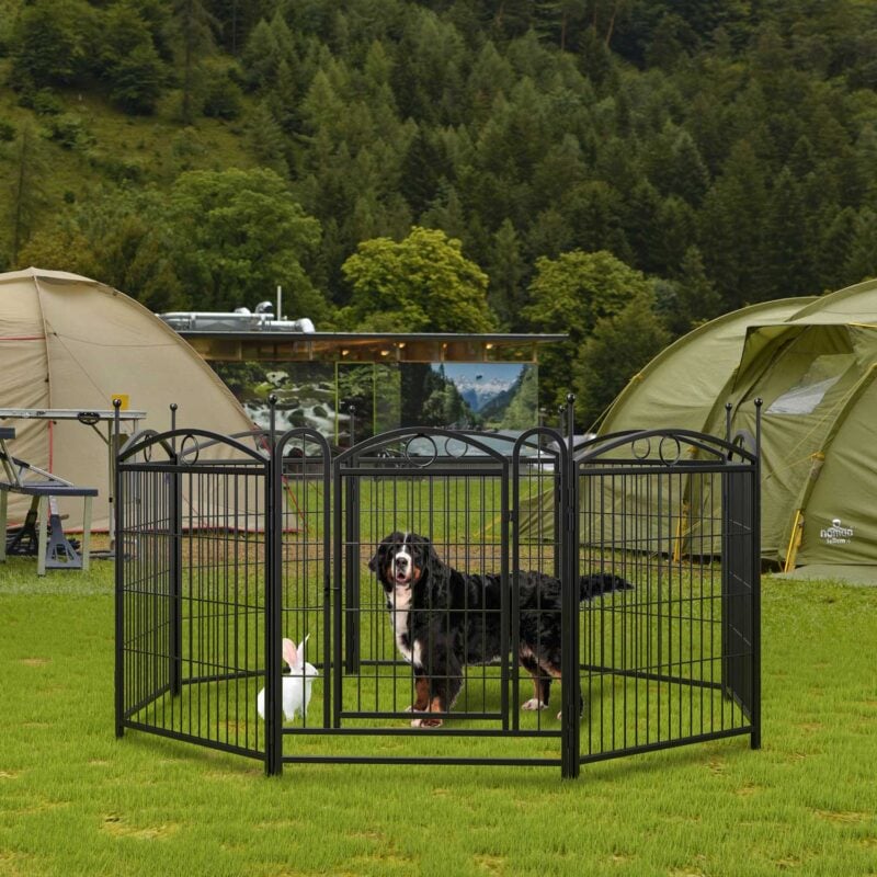 Black metal dog playpen on grass with Bernese Mountain Dog and rabbit, set at a campsite with tents, forest, mountains, and overcast sky in the background.