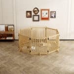 Light wood hexagonal dog playpen with vertical slats, medium dog on mat with bowls, in bright room with herringbone floor and wall art.