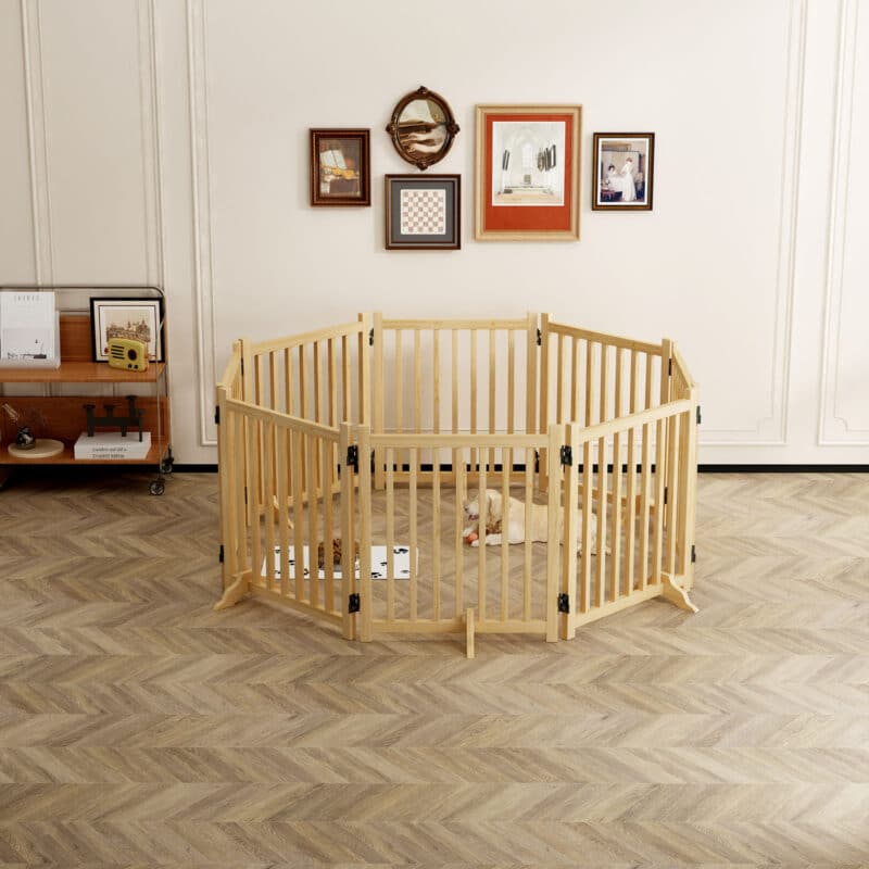 Light wood hexagonal dog playpen with vertical slats, medium dog on mat with bowls, in bright room with herringbone floor and wall art.