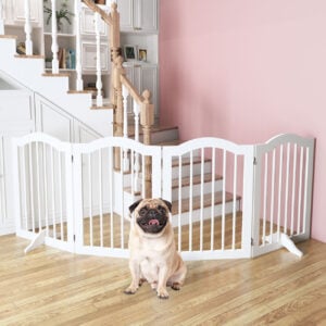 A cheerful Pug rests in front of a white dog gate placed at the base of a wooden staircase, highlighting stair safety.