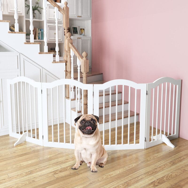 A cheerful Pug rests in front of a white dog gate placed at the base of a wooden staircase, highlighting stair safety.