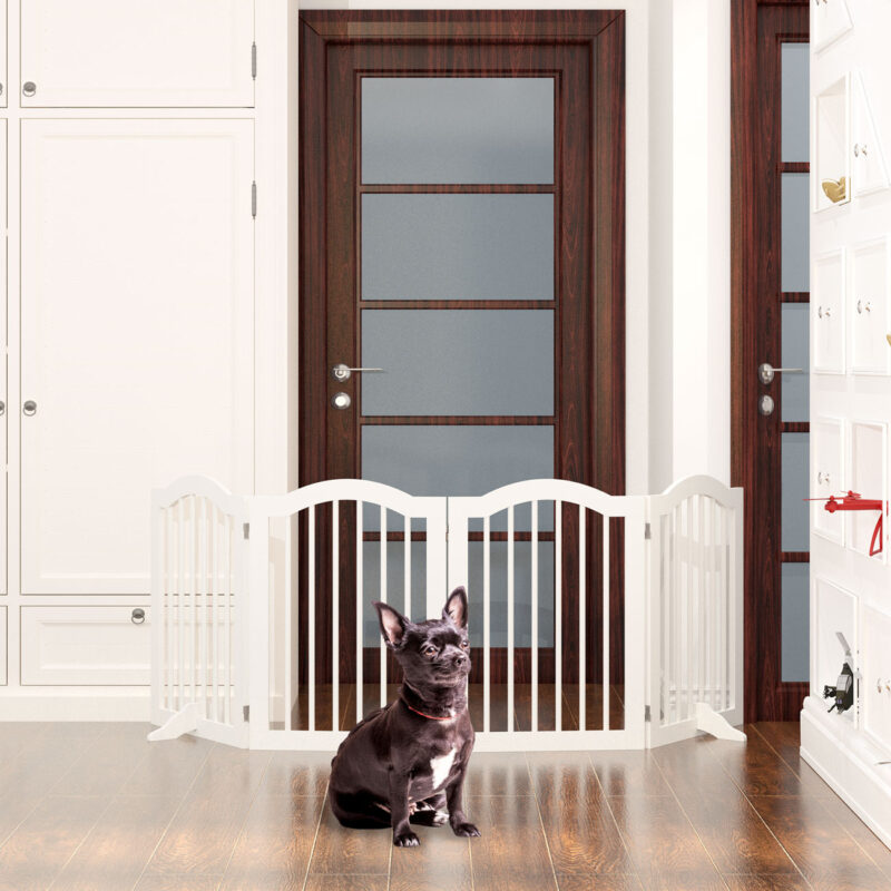 A French Bulldog sits in front of a white freestanding dog barrier in an entryway with dark wood doors.