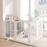 A content Husky puppy lounges behind a freestanding white pet gate in a bright room with large windows, showing an indoor pen setup.